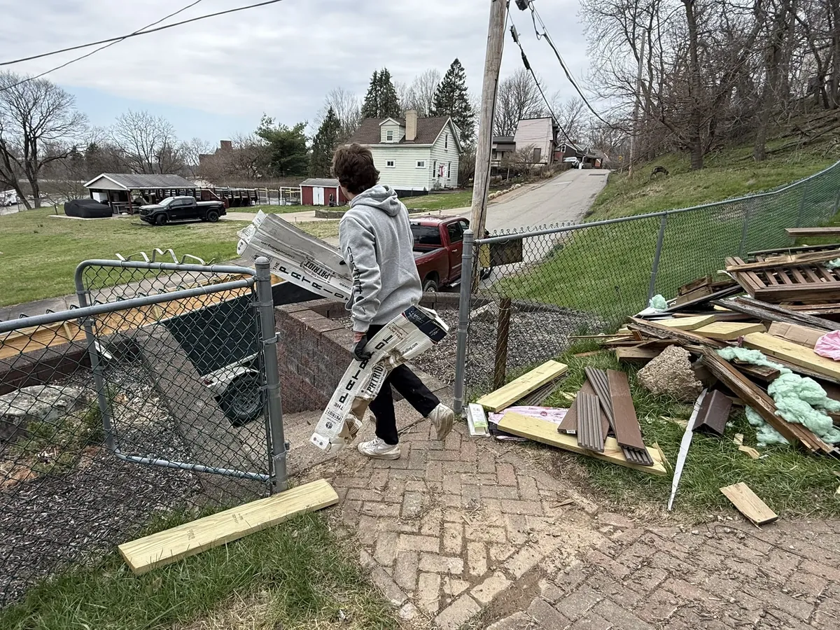 Worker in a gray hoodie carrying old siding past a pile of torn-out lumber and insulation